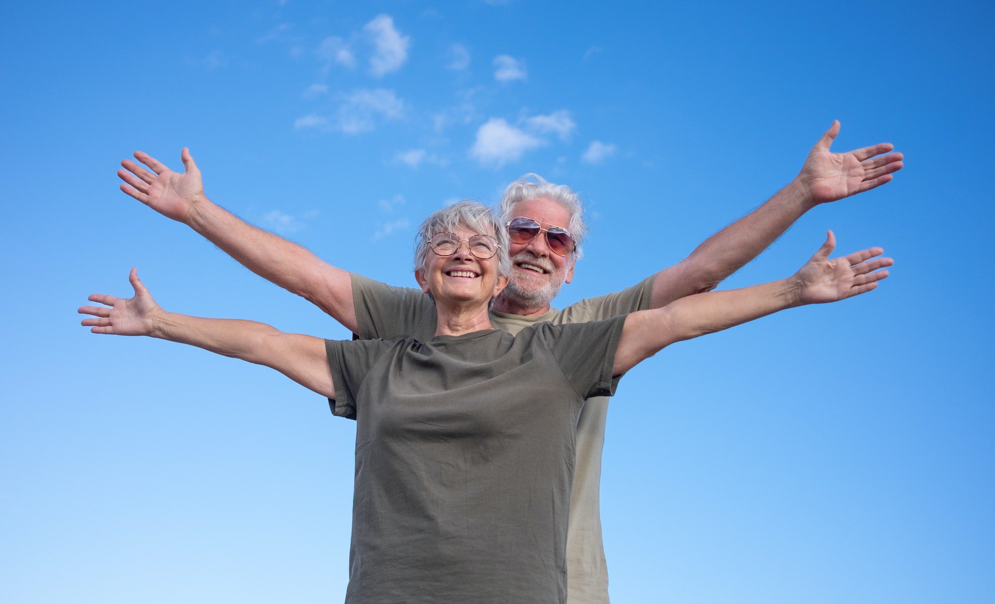 Happy senior couple standing with open arms with blue sky background enjoying outdoor activity as they discuss Bobby Brock Insurance and how they help.